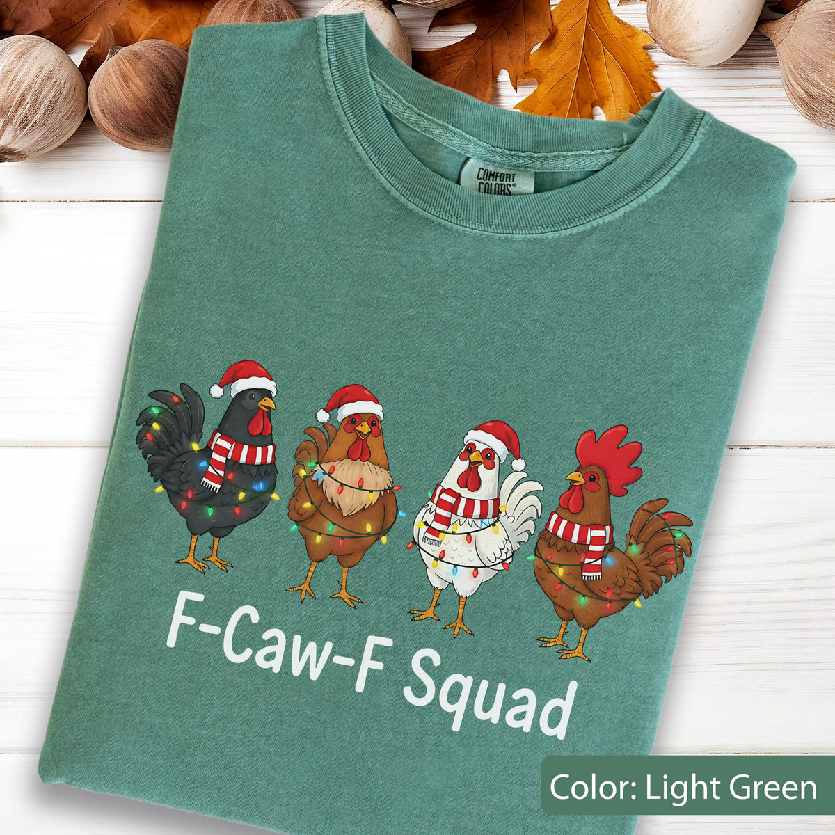 May include: Light green t-shirt featuring four cartoon chickens wearing Santa hats and scarves, adorned with Christmas lights. The text "F-Caw-F Squad" is printed below the chickens. The shirt is a Comfort Colors brand.