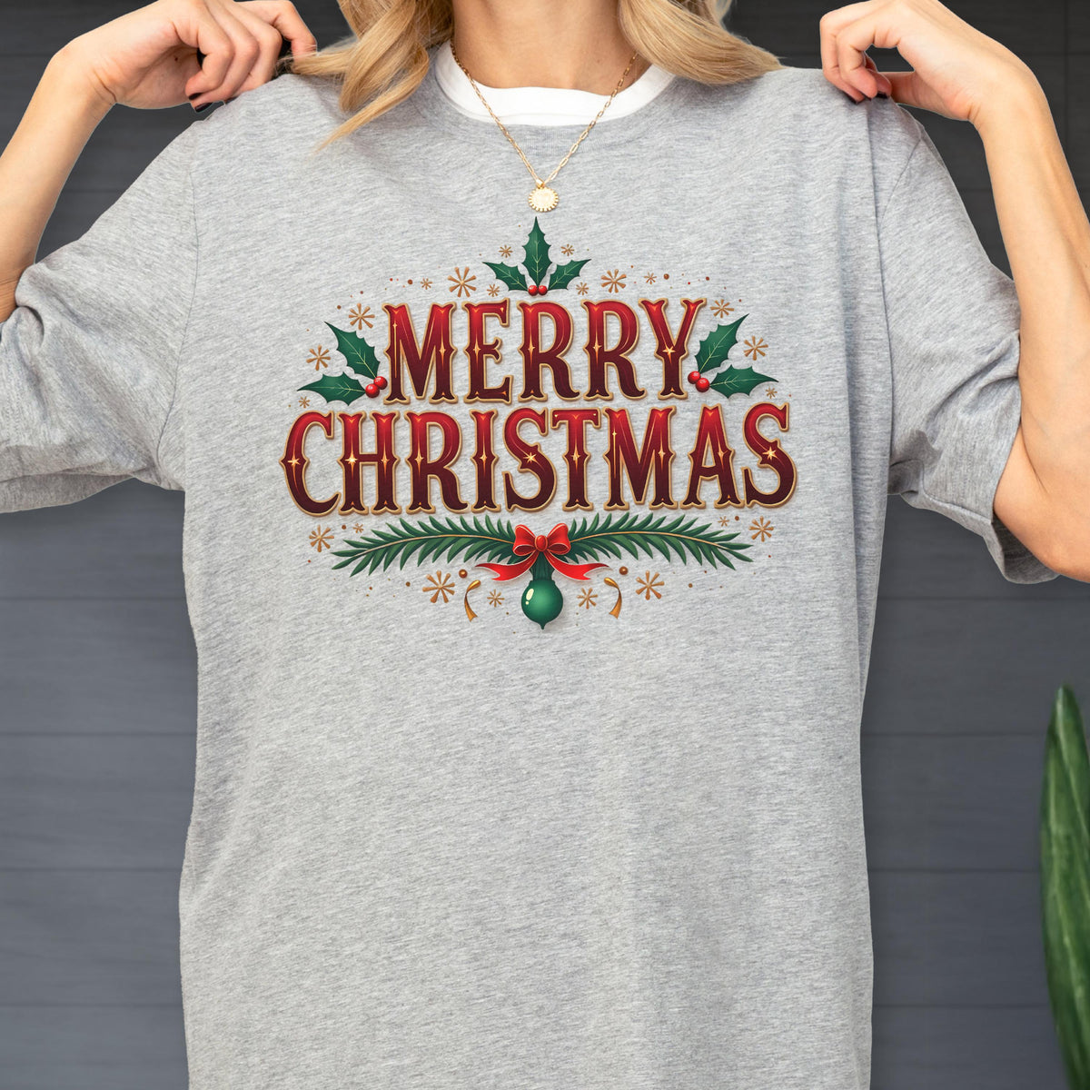May include: A heather gray t-shirt featuring the red and gold text Merry Christmas in a decorative font. The design includes green holly leaves, red berries, pine branches, and a hanging green ornament with a red bow, accented by small gold stars.