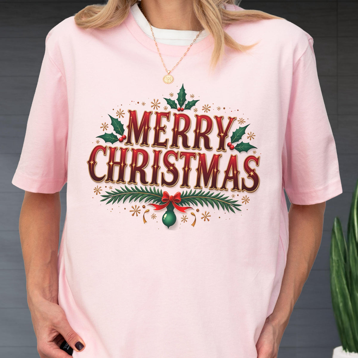 May include: A light pink short-sleeved t-shirt featuring a graphic print with the text Merry Christmas in red and dark brown lettering. The design includes green holly leaves, red berries, pine branches, a red bow, and gold star accents.
