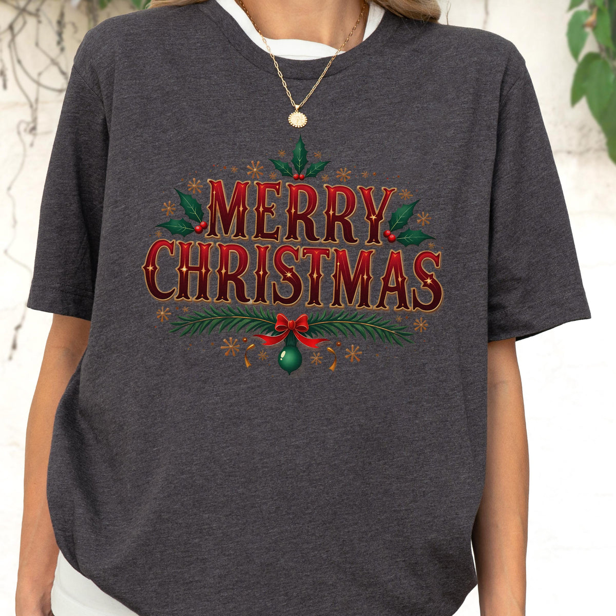 May include: A charcoal gray short-sleeve t-shirt featuring the text Merry Christmas in a bold red font with gold outlines. The design includes green holly leaves, red berries, pine branches, a red bow, and a green ornament with gold snowflakes.