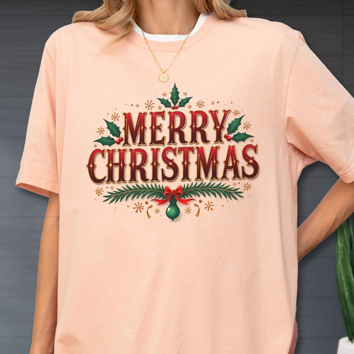 May include: A peach-colored short-sleeve t-shirt featuring a graphic print that reads MERRY CHRISTMAS in bold red and dark brown lettering. The design is decorated with green holly leaves, red berries, pine branches, and a small green ornament.
