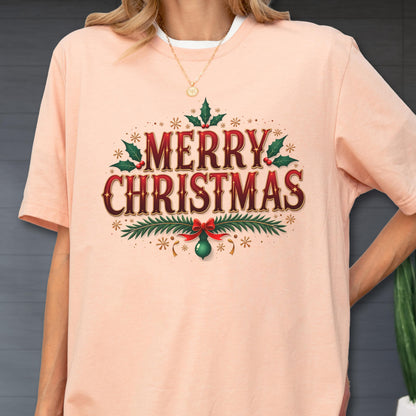 May include: A peach-colored short-sleeve t-shirt featuring a graphic print that reads MERRY CHRISTMAS in bold red and dark brown lettering. The design is decorated with green holly leaves, red berries, pine branches, and a small green ornament.