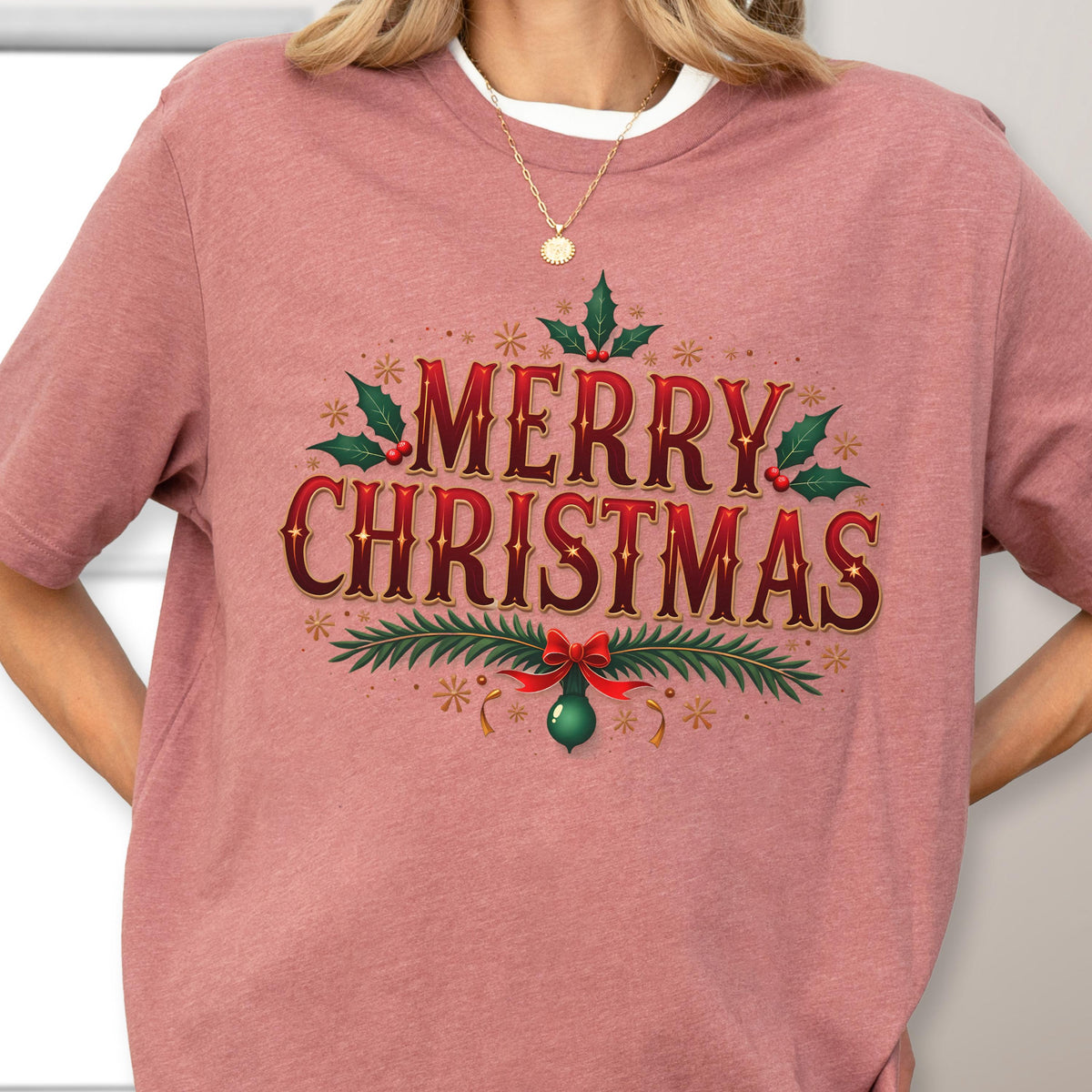 May include: A dusty rose short-sleeved t-shirt featuring a graphic print with the text MERRY CHRISTMAS in dark red lettering. The design includes green holly leaves, red berries, pine branches, a red bow, and small gold star accents.
