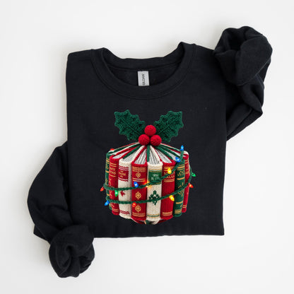 May include: Black sweatshirt featuring a festive design of stacked books, adorned with holly leaves and berries. The books are wrapped with colorful Christmas lights, creating a cozy, holiday-themed look. The sweatshirt is folded.