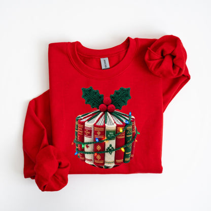 May include: A red sweatshirt featuring a festive design. The design includes a stack of books with a green and red holly and berry accent, wrapped with a string of colorful lights. The sweatshirt has long sleeves.