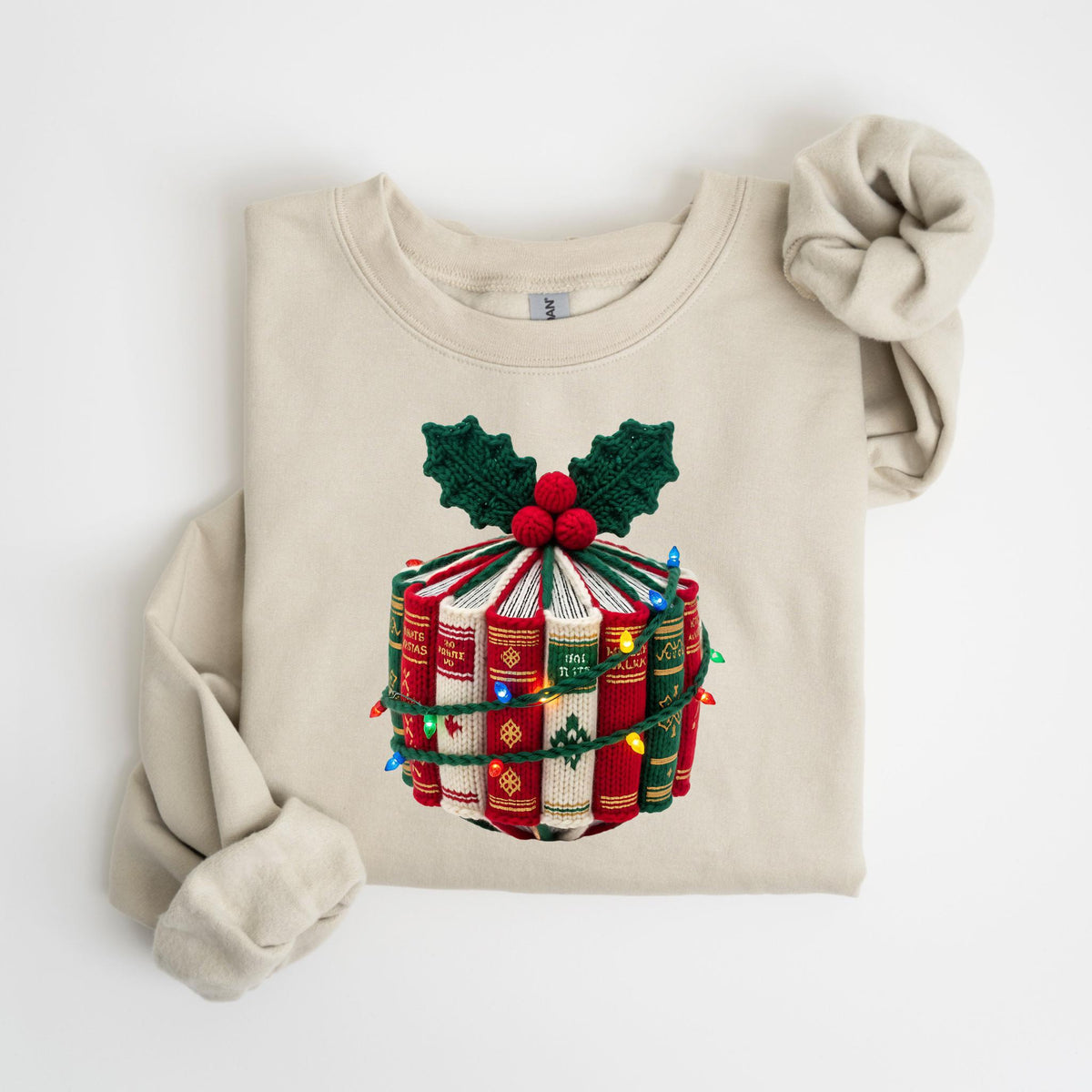 May include: Beige sweatshirt with a festive design featuring a stack of books wrapped with green and red yarn, topped with holly and berries. The books are adorned with a string of colorful lights, creating a cozy, holiday-themed look.