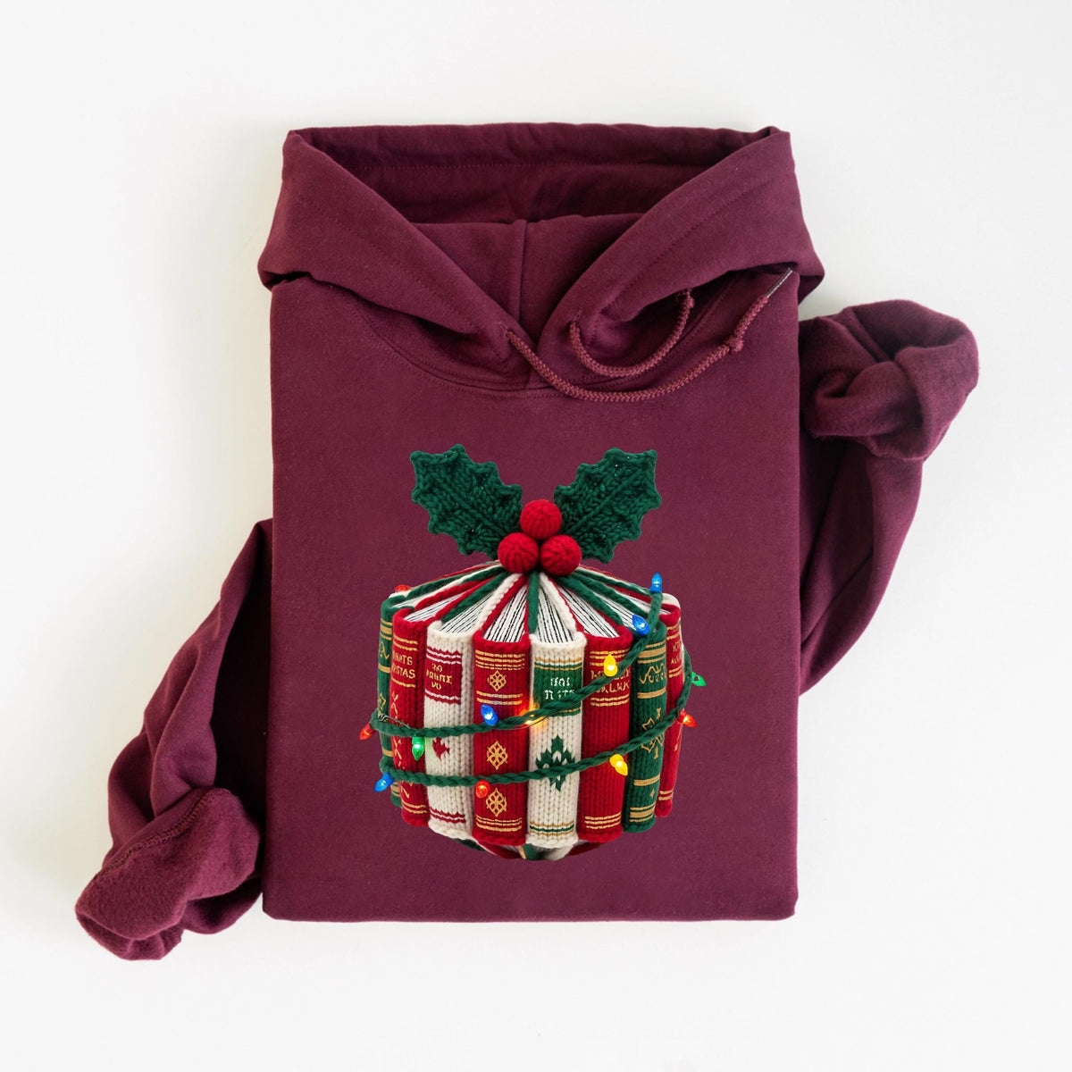 May include: Burgundy hoodie with a festive design. The design features a stack of books wrapped with string lights and topped with holly and berries. The books are red, green, and white.