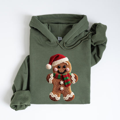 May include: An olive green hooded sweatshirt featuring a gingerbread man design. The gingerbread man wears a red Santa hat and a red and green scarf. The design is centered on the front of the sweatshirt.