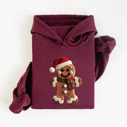 May include: Burgundy hoodie featuring a gingerbread man design. The gingerbread man wears a Santa hat and a scarf with red and green stripes. The hoodie has a drawstring hood and long sleeves. The gingerbread man has a smiling face and two white snowflakes.