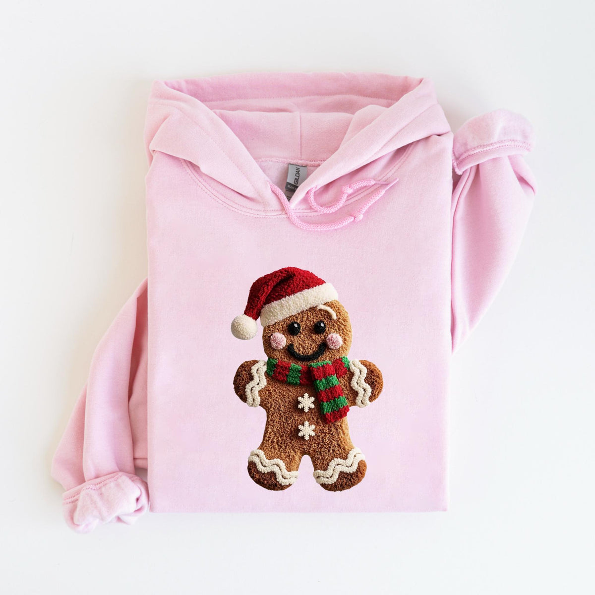May include: A light pink hoodie featuring a gingerbread man design. The gingerbread man wears a red Santa hat and a green and red striped scarf. The gingerbread man has white icing details and two white snowflakes.