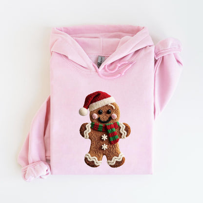 May include: A light pink hoodie featuring a gingerbread man design. The gingerbread man wears a red Santa hat and a green and red striped scarf. The gingerbread man has white icing details and two white snowflakes.