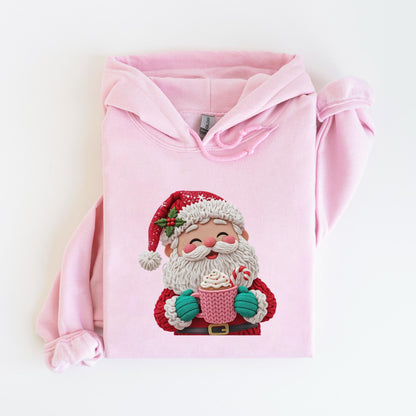 May include: A light pink hoodie featuring a Santa Claus graphic. The Santa illustration shows a smiling face, a red hat, and a white beard, holding a pink mug with whipped cream and a candy cane. The hoodie has a drawstring hood and long sleeves.