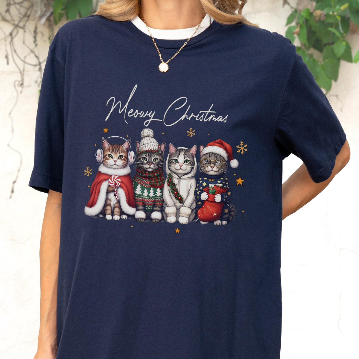 May include: A navy blue t-shirt featuring a graphic of four cats dressed in winter clothing with the text Meowy Christmas. The cats wear items including a red cape, a knit sweater, a white hoodie, and a Santa hat, surrounded by gold snowflakes.