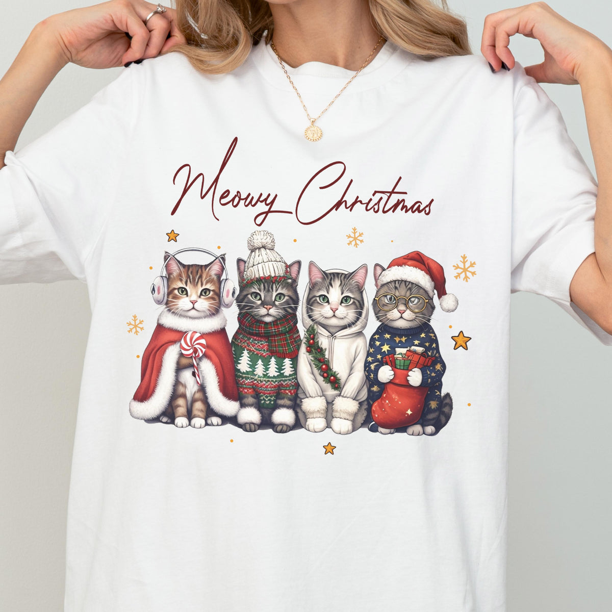 May include: A white short-sleeve t-shirt featuring a graphic of four cats dressed in winter holiday attire with the text Meowy Christmas. The cats wear earmuffs, a knit hat, a hoodie, and a Santa hat, surrounded by small gold stars and snowflakes.