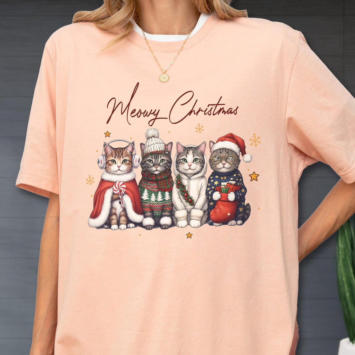 May include: A peach-colored t-shirt featuring a graphic of four cats dressed in winter holiday attire with the text Meowy Christmas. The cats wear accessories including earmuffs, a knit hat, a scarf, and a Santa hat, surrounded by small stars.