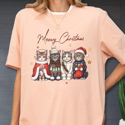 May include: A peach-colored t-shirt featuring a graphic of four cats dressed in winter holiday attire with the text Meowy Christmas. The cats wear accessories including earmuffs, a knit hat, a scarf, and a Santa hat, surrounded by small stars.