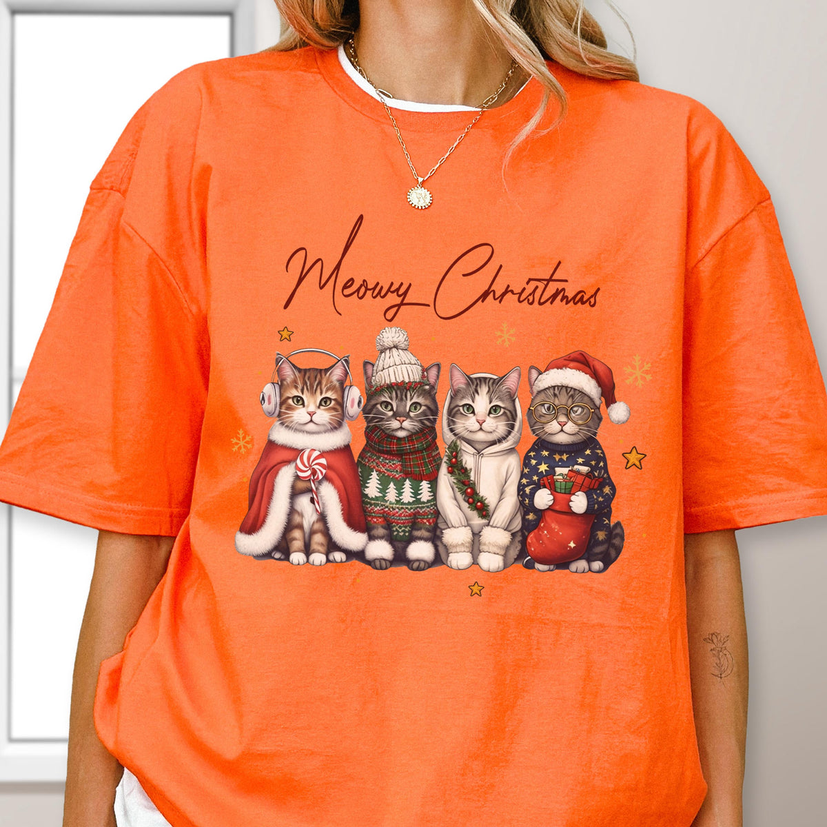 May include: An orange short-sleeve t-shirt featuring a graphic of four cats dressed in winter holiday attire. The text "Meowy Christmas" is printed above the cats, who are wearing earmuffs, a knit hat, a hoodie, and a Santa hat with a stocking.