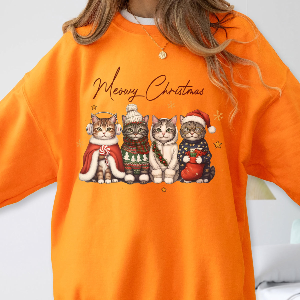 May include: An orange crewneck sweatshirt featuring a graphic of four cats wearing winter accessories and the text Meowy Christmas. The cats are dressed in a red cape with earmuffs, a knit hat and scarf, a white hoodie, and a Santa hat with a sweater.