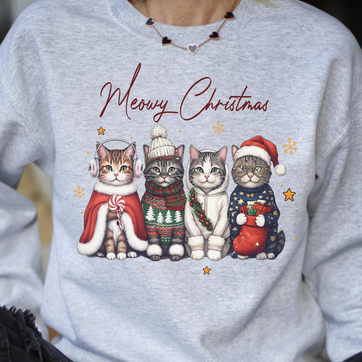 May include: A light gray sweatshirt featuring the text Meowy Christmas in dark red script. Below the text are four illustrated cats wearing winter accessories, including earmuffs, a knit hat, a hoodie, and a Santa hat, with small gold stars.