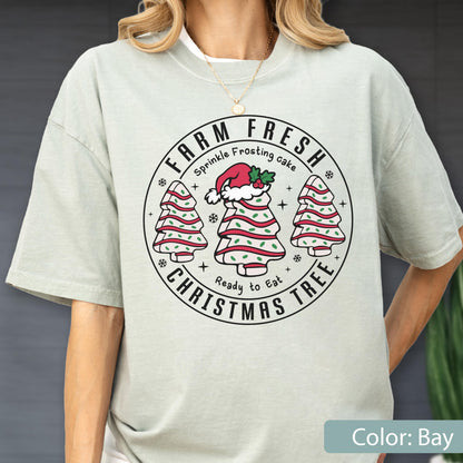 May include: A bay-colored t-shirt featuring a circular graphic with the text FARM FRESH CHRISTMAS TREE. The design includes three tree-shaped cakes with red frosting stripes and green sprinkles, with the center cake wearing a Santa hat.