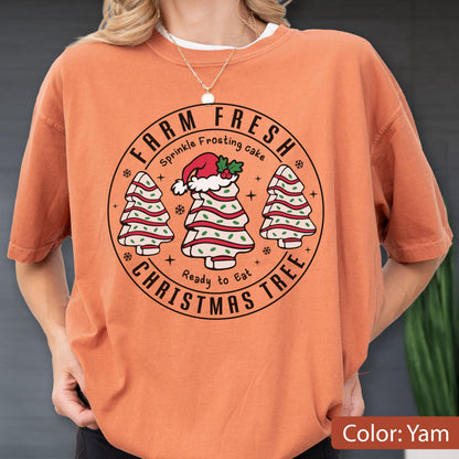 May include: A yam-colored short-sleeve t-shirt featuring a circular graphic with the text "FARM FRESH CHRISTMAS TREE" and "Sprinkle Frosting cake Ready to Eat". The design includes three stylized cake trees, one wearing a Santa hat.