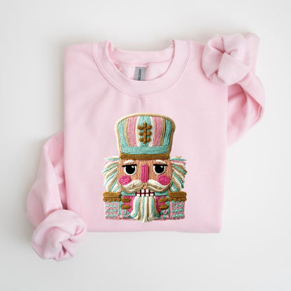 May include: A light pink sweatshirt featuring a detailed embroidered nutcracker design. The nutcracker has a teal, pink, and gold color scheme, with a large hat and a detailed face. The sweatshirt is a crew neck style.