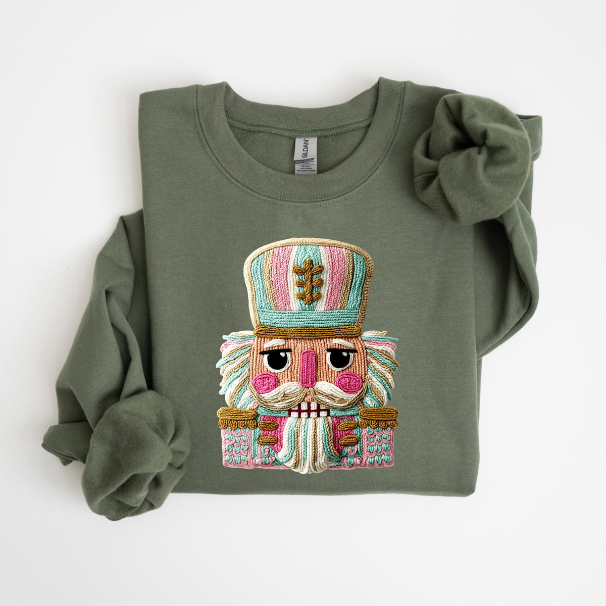 May include: Olive green sweatshirt featuring a colorful embroidered nutcracker design. The nutcracker has a detailed face, mustache, and a hat with pink, blue, and gold accents. A festive and cozy apparel item.