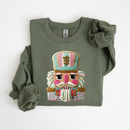 May include: Olive green sweatshirt featuring a colorful embroidered nutcracker design. The nutcracker has a detailed face, mustache, and a hat with pink, blue, and gold accents. A festive and cozy apparel item.