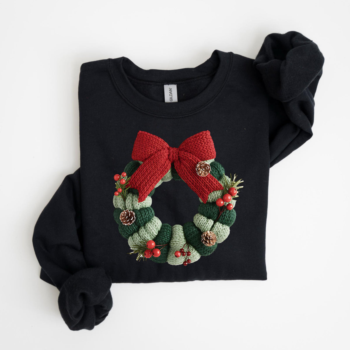 May include: Black sweatshirt featuring a knitted Christmas wreath design. The wreath is composed of green and light green sections, adorned with a red bow, berries, and pinecones. The long sleeves are folded.
