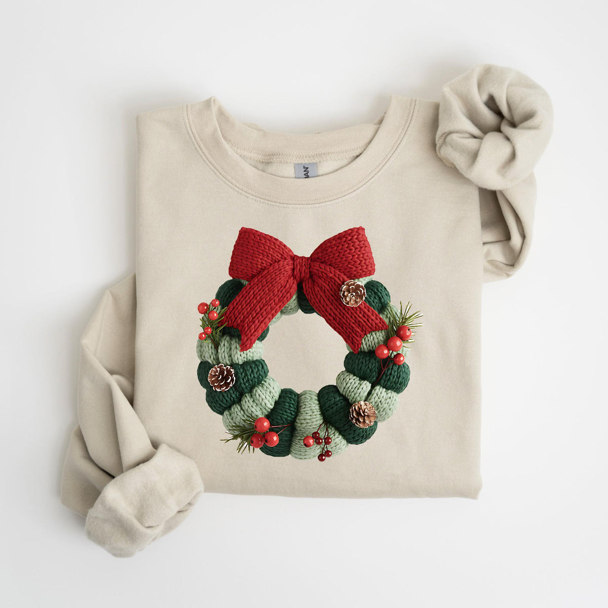 May include: A beige sweatshirt featuring a knitted Christmas wreath design. The wreath is composed of green and light green sections, adorned with red berries, pinecones, and a large red bow at the top. The sleeves are folded.