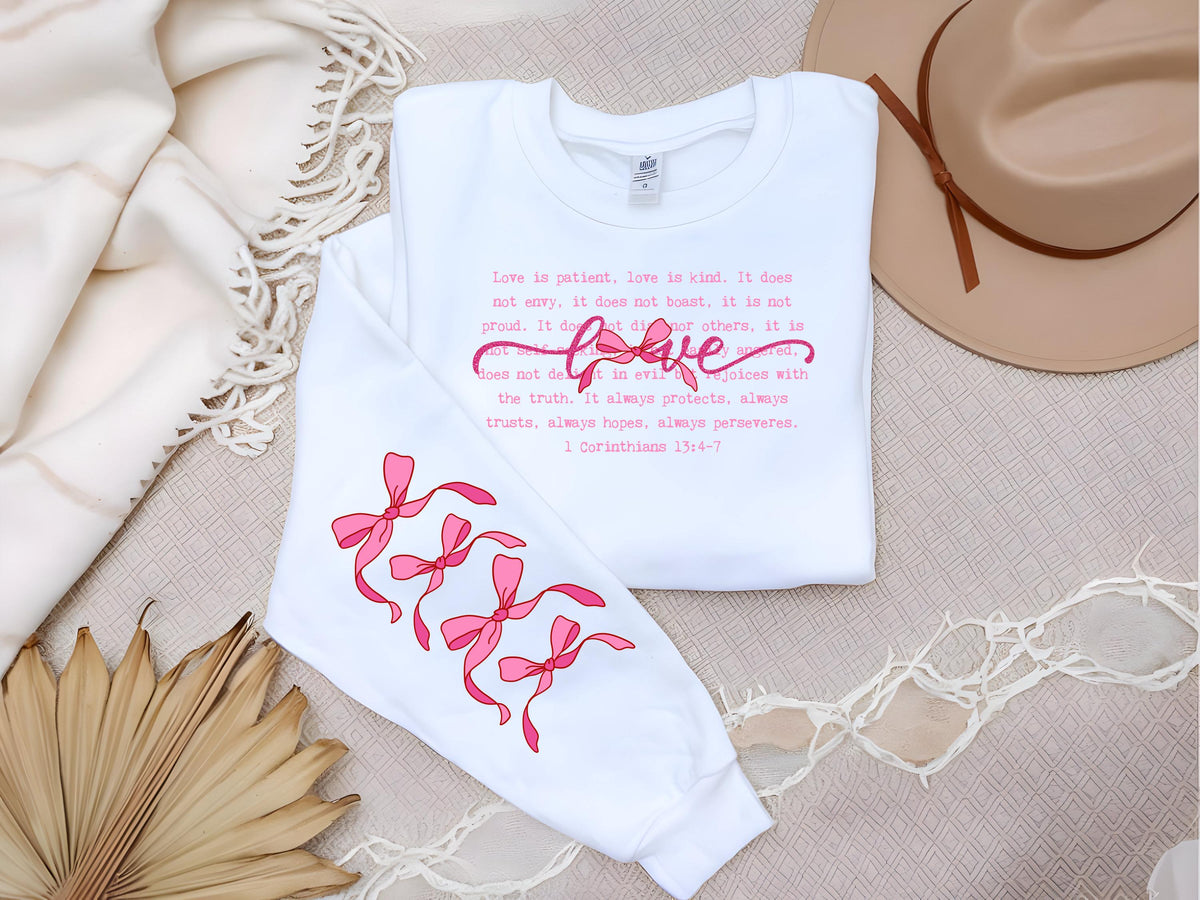 May include: White long-sleeved shirt with pink text and bow graphics. The text includes the word "love" in a cursive font and a Bible verse. The sleeves feature three pink bow designs.