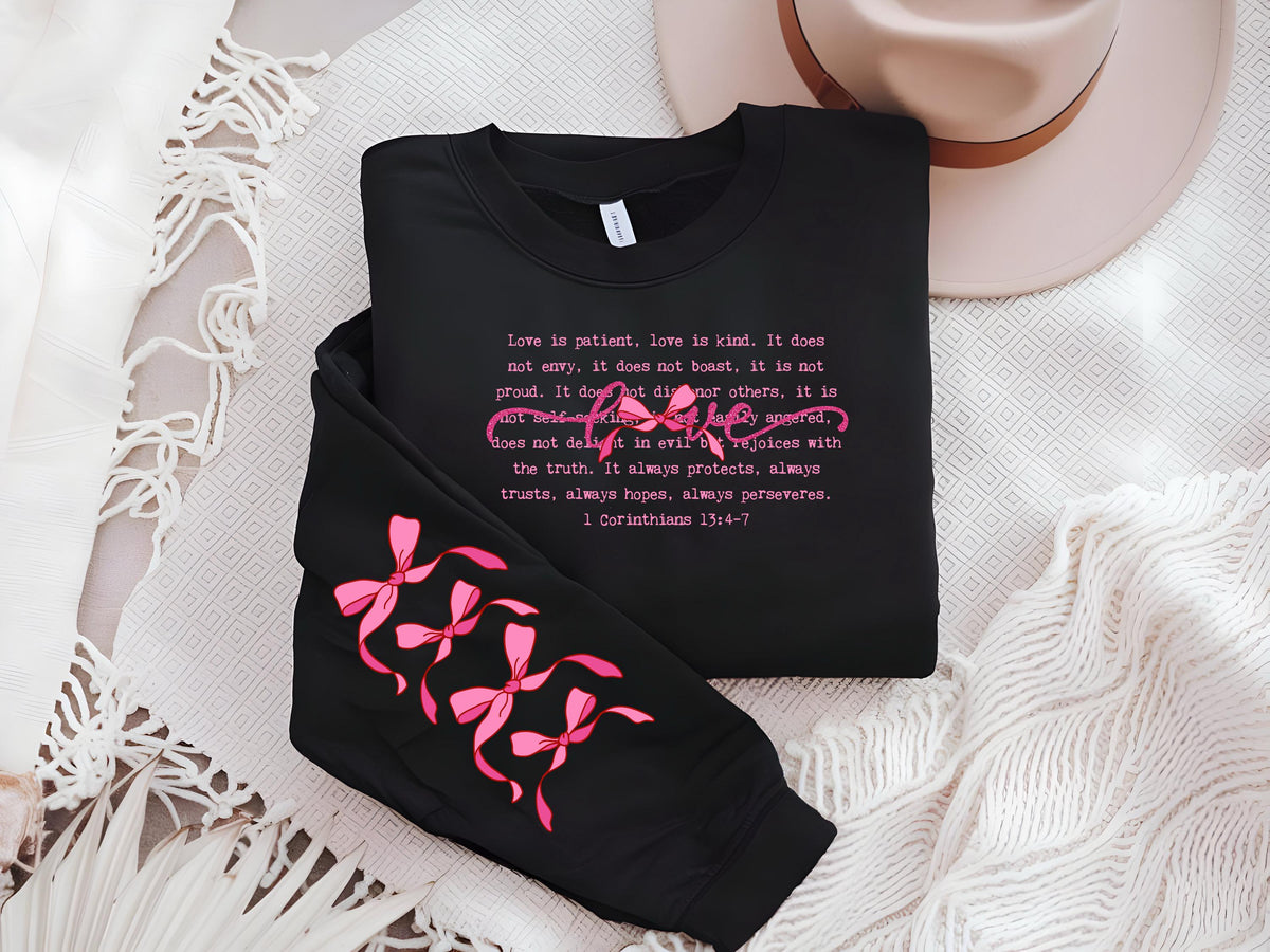 May include: Black sweatshirt with pink text and bow accents. The text reads "Love is patient, love is kind" from 1 Corinthians 13:4-7. The sleeve features pink bow designs. A neutral-toned hat is in the background.
