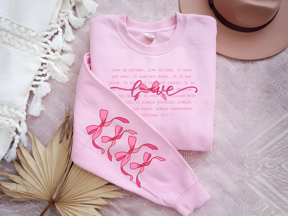 May include: A light pink sweatshirt with the word "love" in cursive and the text of 1 Corinthians 13:4-7. The sleeve features a design of pink bows. The sweatshirt is a casual, comfortable garment.