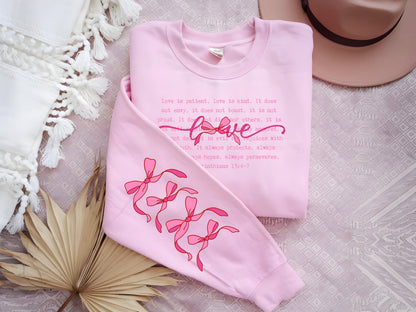May include: A light pink sweatshirt with the word "love" in cursive and the text of 1 Corinthians 13:4-7. The sleeve features a design of pink bows. The sweatshirt is a casual, comfortable garment.