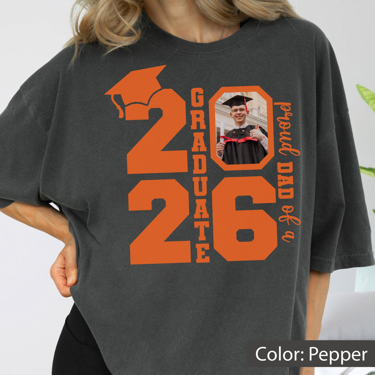 May include: Pepper-colored t-shirt with orange graphics. The design includes the number "26" with a graduation cap, the word "GRADUATE", and a photo of a graduate. The text "proud DAD of a" is also present.
