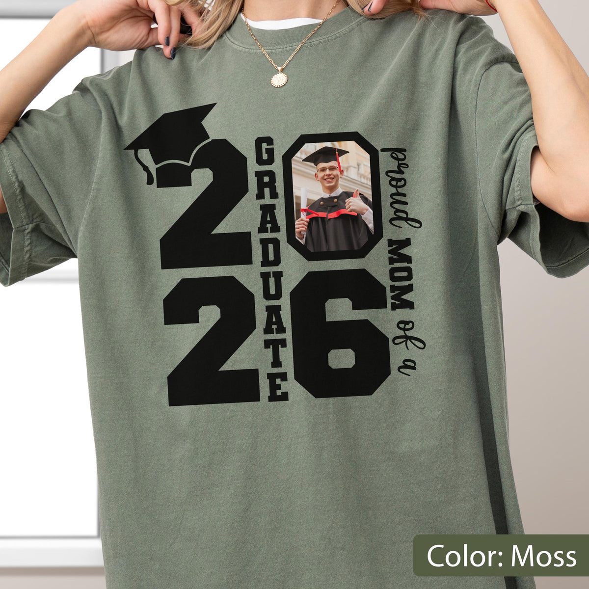 May include: Moss green t-shirt with a graduation theme. The shirt features the number "26" with a graduation cap graphic, the word "GRADUATE", a photo, and the text "proud MOM of a".