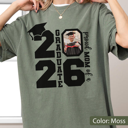 May include: Moss green t-shirt with a graduation theme. The shirt features the number "26" with a graduation cap graphic, the word "GRADUATE", a photo, and the text "proud MOM of a".