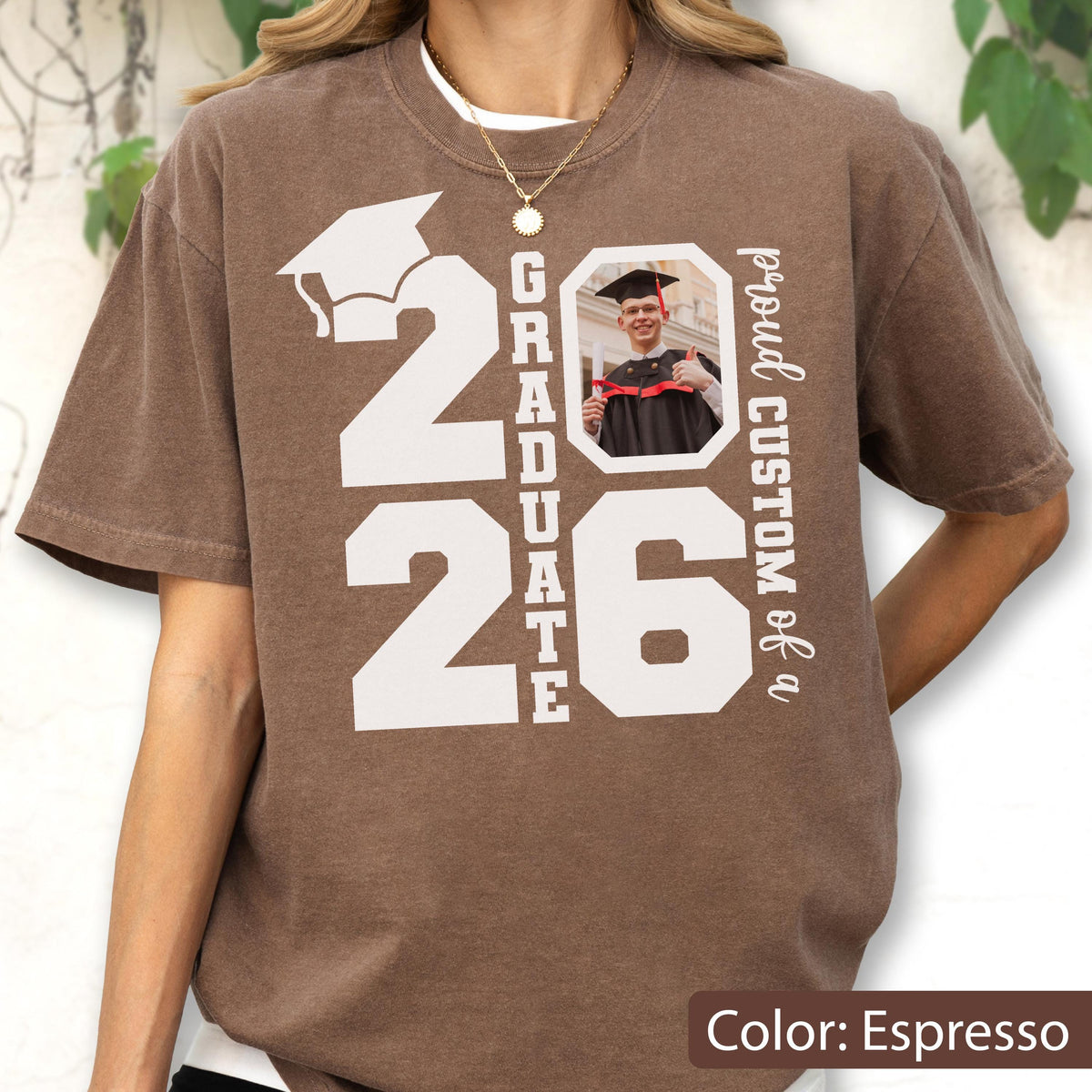 May include: Espresso-colored t-shirt with a graduation theme. The design includes the year "2026", a graduation cap, the word "GRADUATE", and a photo. The text "proud CUSTOM of a" is also visible.