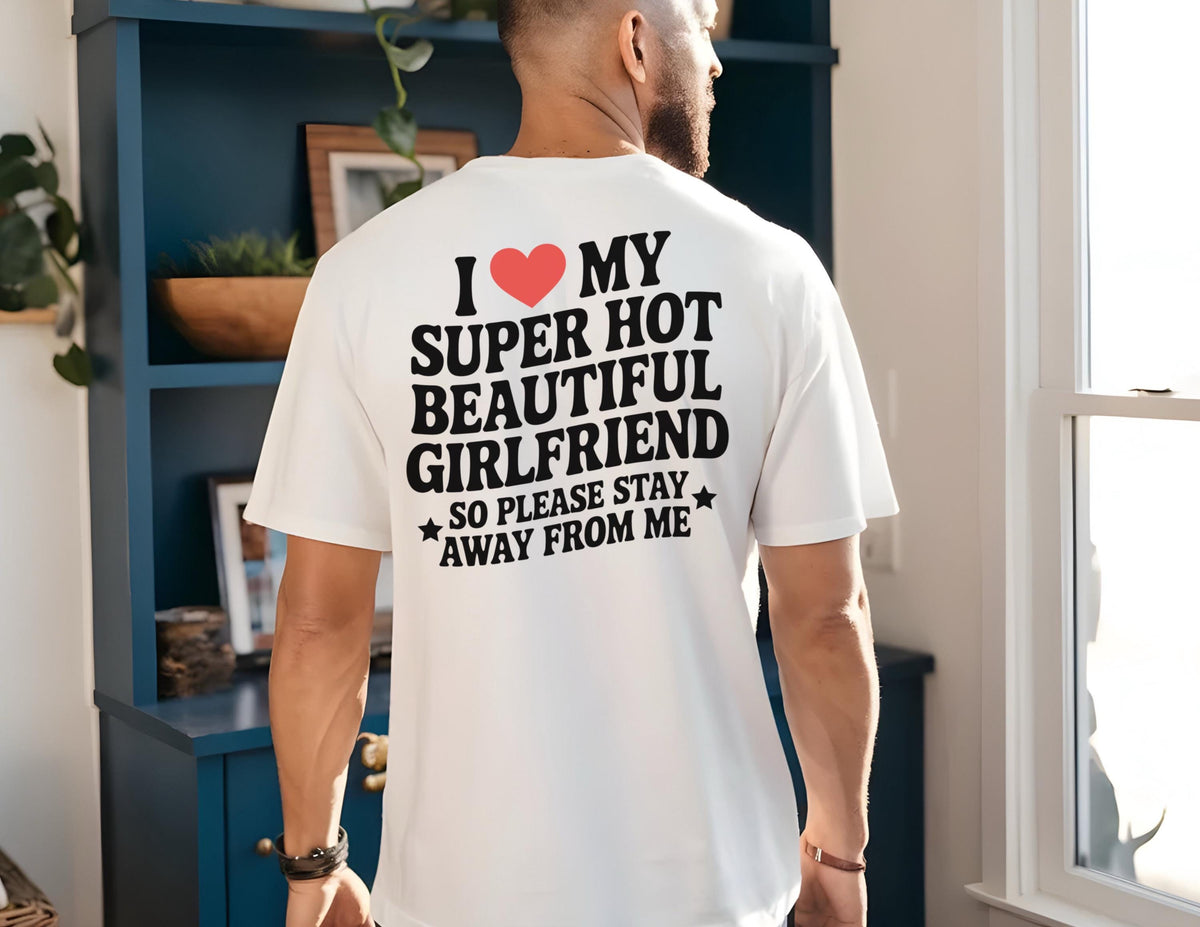 May include: White t-shirt with black text and a red heart graphic. The text reads "I ❤️ MY SUPER HOT BEAUTIFUL GIRLFRIEND SO PLEASE STAY AWAY FROM ME". The shirt is worn by a person.
