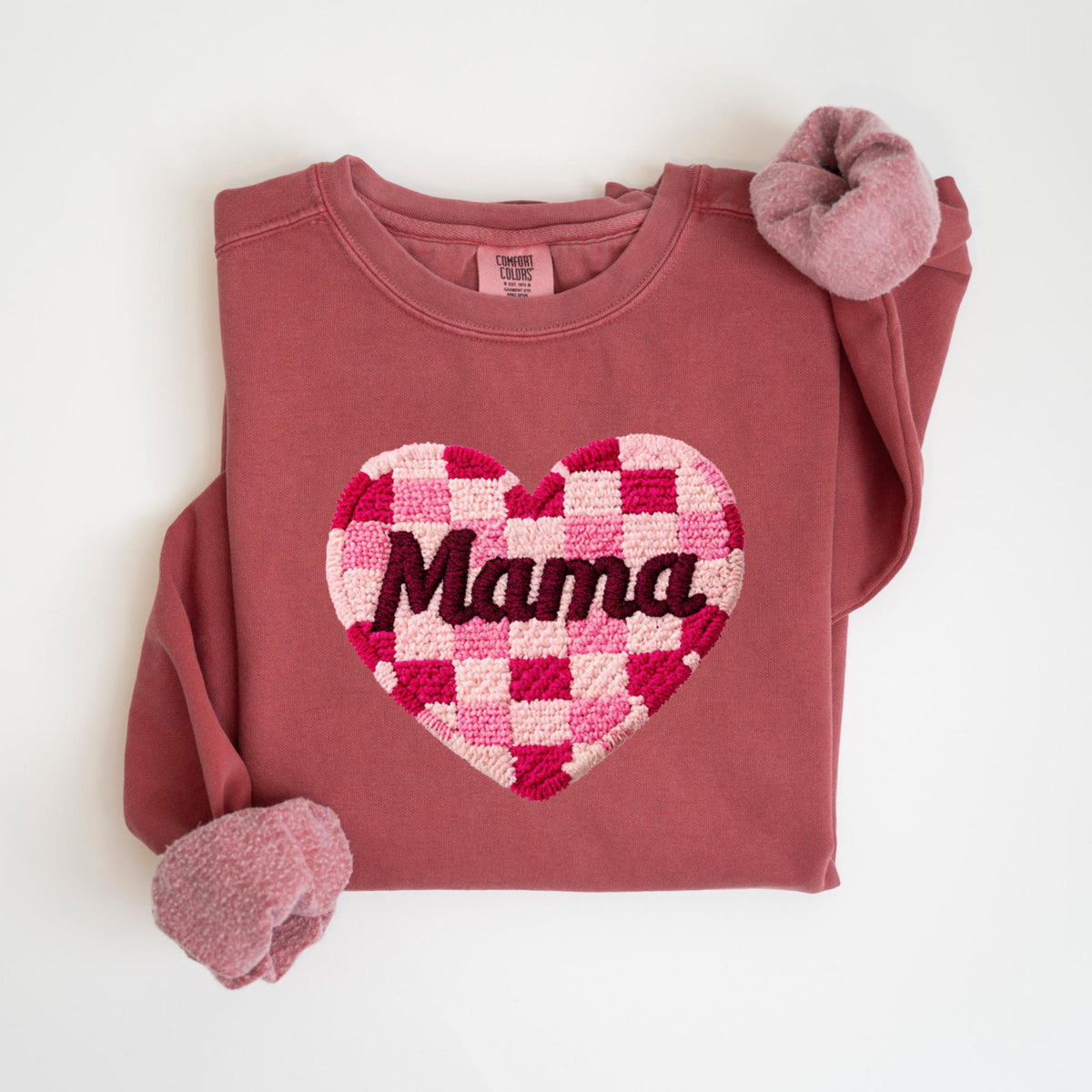 May include: A mauve-colored sweatshirt with a large heart-shaped patch. The patch features a checkerboard pattern in shades of pink and red, with the word "Mama" embroidered in dark brown.