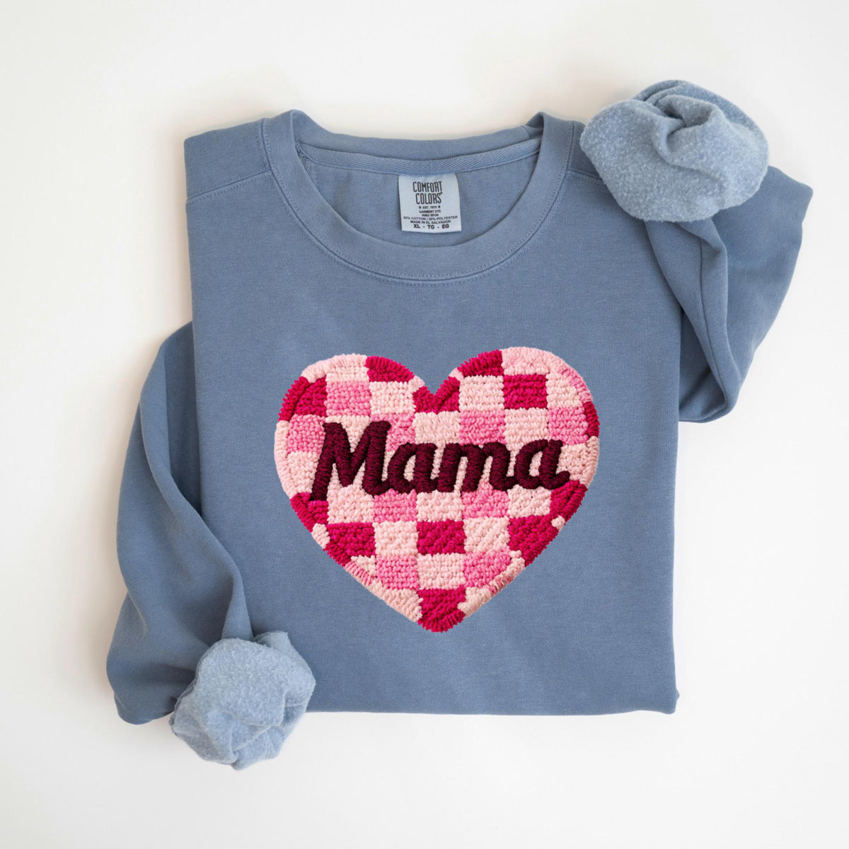 May include: A blue crewneck sweatshirt with a large, embroidered heart. The heart features a pink and red checkerboard pattern with the word "Mama" in a dark red script. The sweatshirt has long sleeves and a soft, comfortable appearance.
