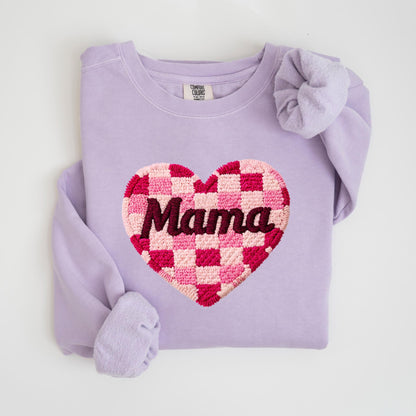 May include: A light purple sweatshirt with a large heart design. The heart is a checkerboard pattern in shades of pink and red, with the word "Mama" embroidered in a dark red color. The sweatshirt has long sleeves and a crew neck.