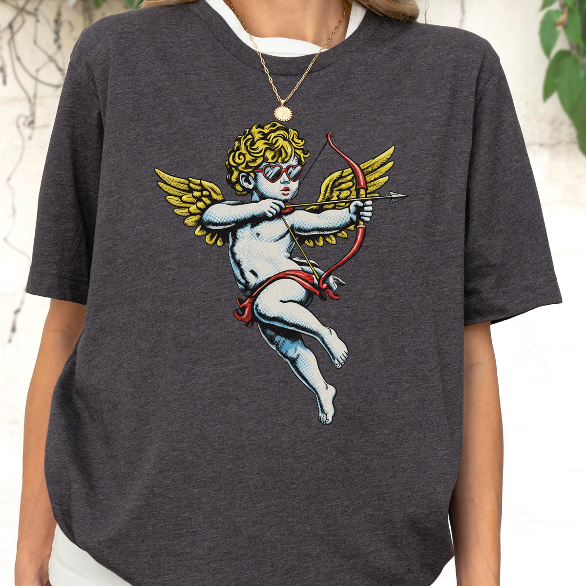 May include: A dark charcoal gray short-sleeve t-shirt featuring a graphic print of a cherub with curly blonde hair, wearing red heart-shaped sunglasses, and holding a red bow and arrow. The cherub has large yellow wings and is depicted in flight.