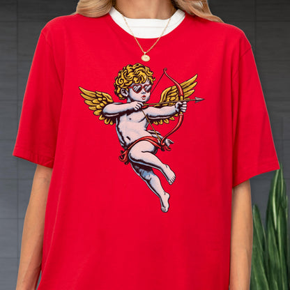 May include: A bright red short-sleeved t-shirt featuring a graphic of a cherub with curly blonde hair, wearing heart-shaped sunglasses, and holding a red bow and arrow. The cherub has large golden wings and is positioned in the center of the shirt.