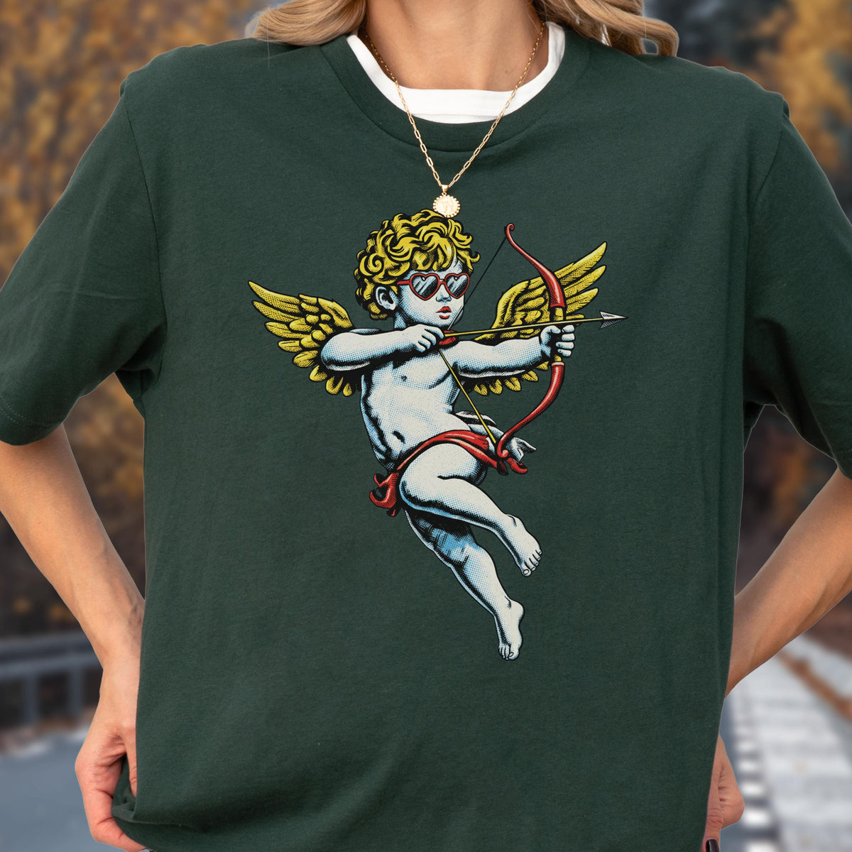 May include: A dark green short-sleeve t-shirt featuring a graphic of a cherub with curly blonde hair, wearing red heart-shaped sunglasses, and holding a red bow and arrow with yellow wings. The shirt has a crew neckline and a white undershirt layer.