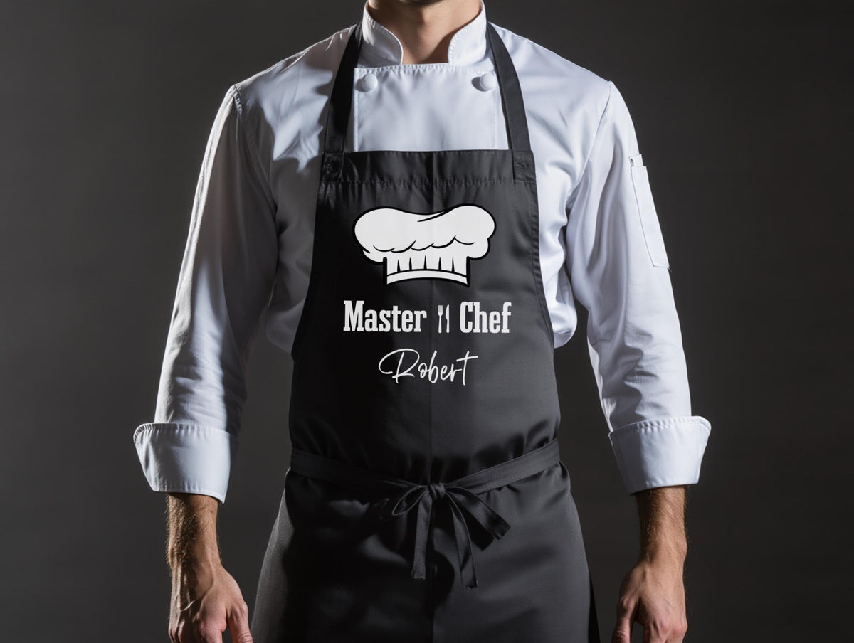 May include: Black apron with white text and a chef's hat graphic. The text reads "Master Chef" above the name "Robert". The apron is tied at the waist and has black straps. The person is wearing a white chef's coat.