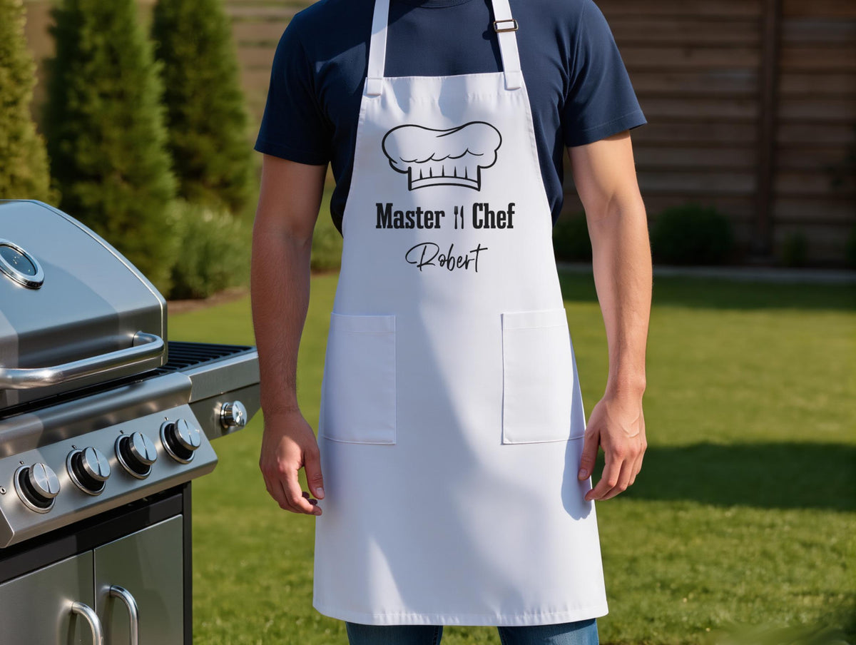 May include: White apron with two pockets and a chef hat graphic. The text "Master Chef" and a name are printed in black. The apron is worn in front of a stainless steel grill.