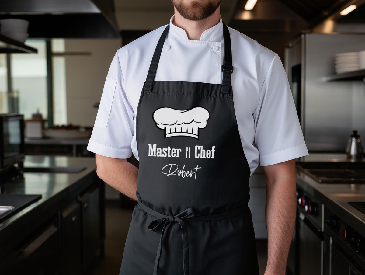 May include: Black apron with a white chef hat graphic and the text "Master Chef Robert". The apron is worn over a white chef's coat. The apron has a neck strap and waist ties.