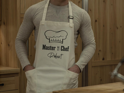 May include: Cream-colored apron with the text "Master Chef" and a chef's hat graphic in black. The name "Robert" is printed below. The apron has two front pockets and a neck strap. The person is wearing a long-sleeved gray shirt.