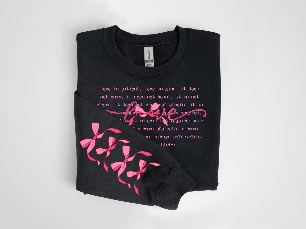 May include: Black sweatshirt with pink text and bow accents. The text reads "Love is patient, love is kind..." with the word "Love" in a cursive font. Pink bows are on the left sleeve. The sweatshirt is folded.